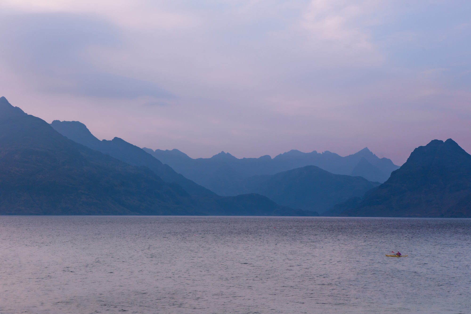 A kayaker in a yellow boat on still water below the blue ridges of the Cuillin at dusk